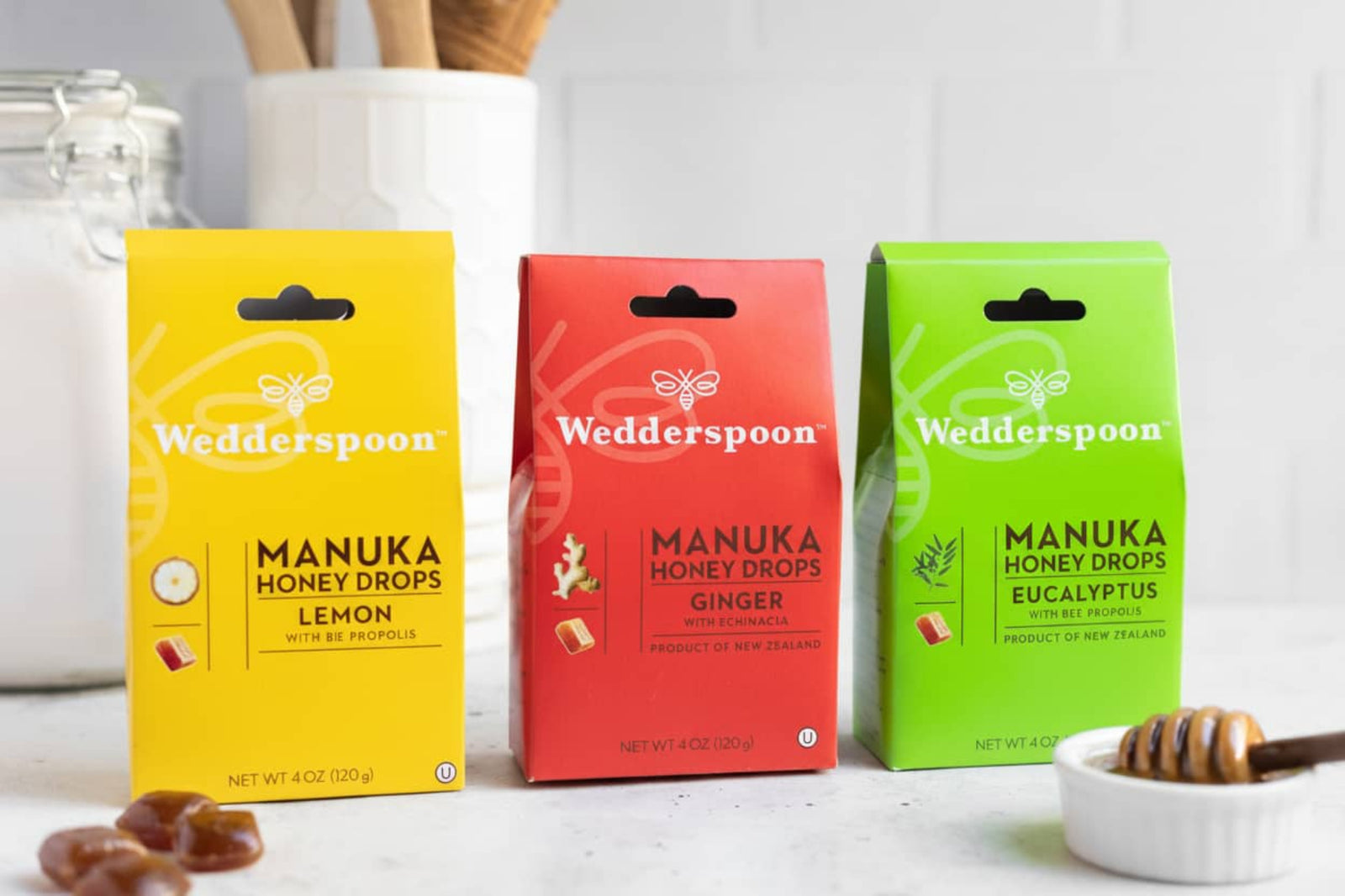 Three boxes of Wedderspoon Manuka Honey Drops on a white surface with honey and honeycombs. Ginger, Lemon and Eucalyptus Flavour