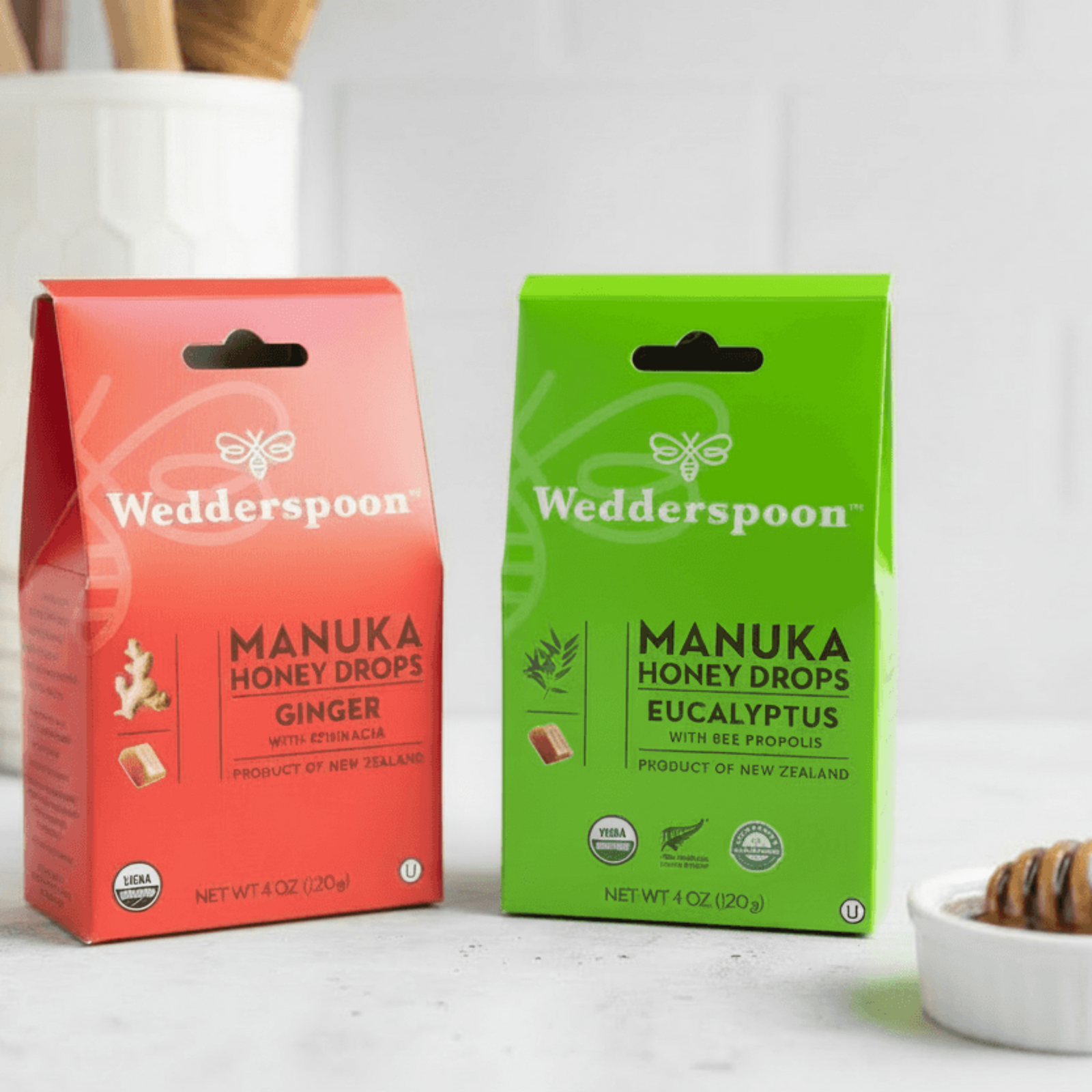 Two Wedderspoon Manuka Honey Drop packages, one Ginger and one Eucalyptus, on a white surface with honeycomb and jar in the background.