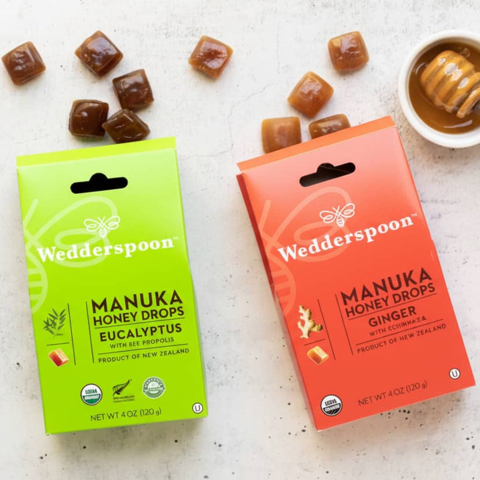Two boxes of Wedderspoon Manuka Honey Drops on a light surface with honey and honeycomb - Eucalyptus and Ginger Flavour