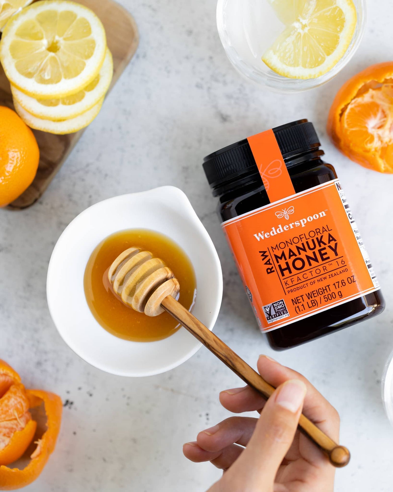 Jar of Wedderspoon raw Manuka honey KFactor 16 with honey dipper, fresh citrus slices surrounding