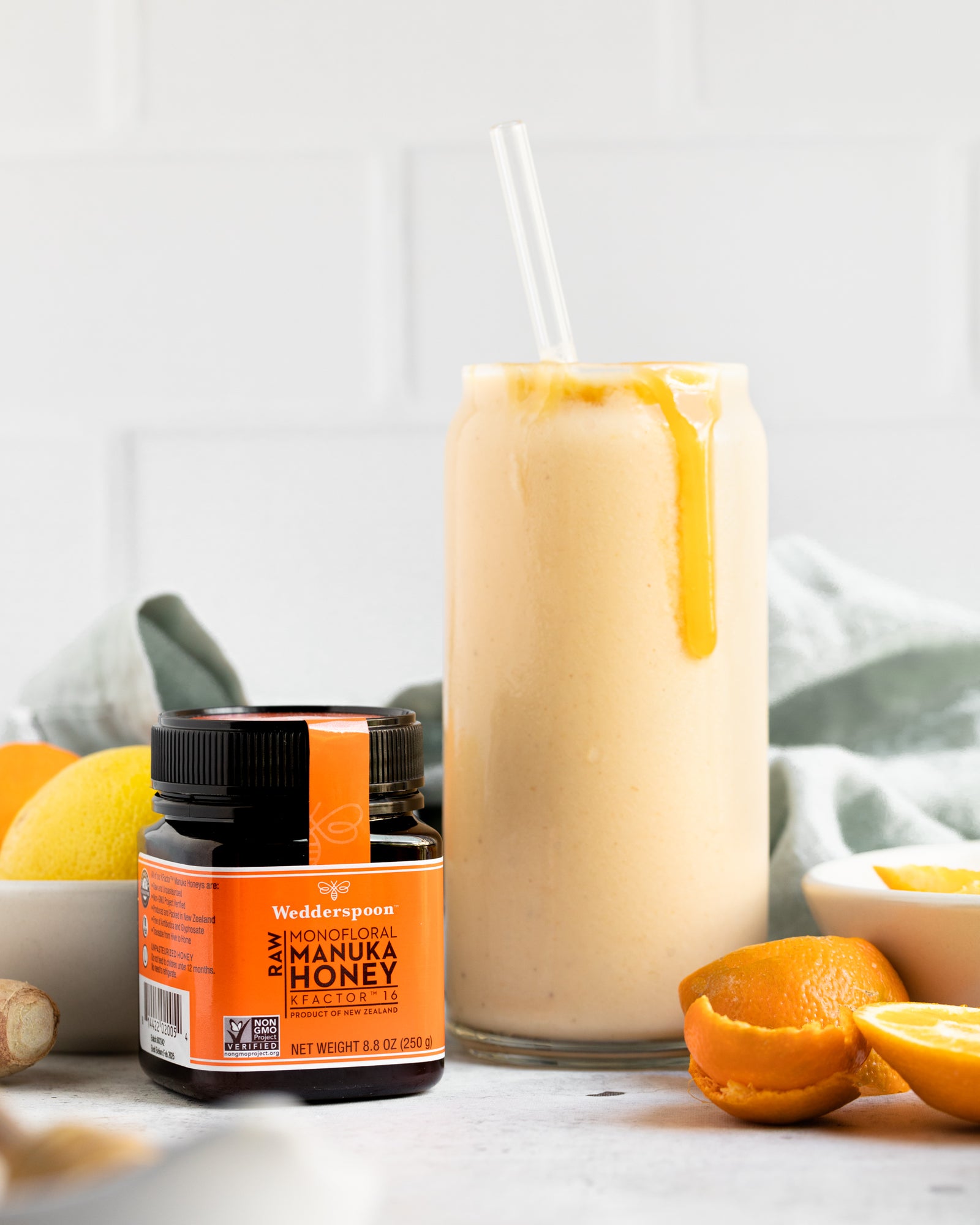 Wedderspoon raw Manuka honey KFactor 16+ jar beside citrus smoothie with honey drizzle
