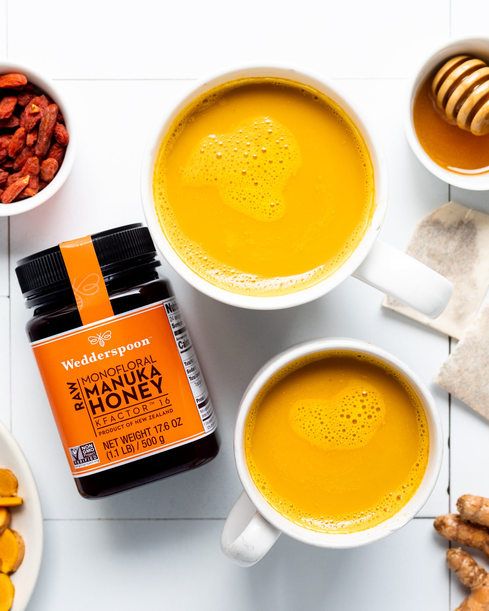 Wedderspoon raw Manuka honey KFactor 16 jar next to two cups of golden tea with honey dipper and natural ingredients