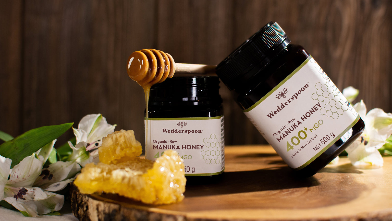 Bottles of Manuka honey on a wooden surface with flowers and a honey dipper.