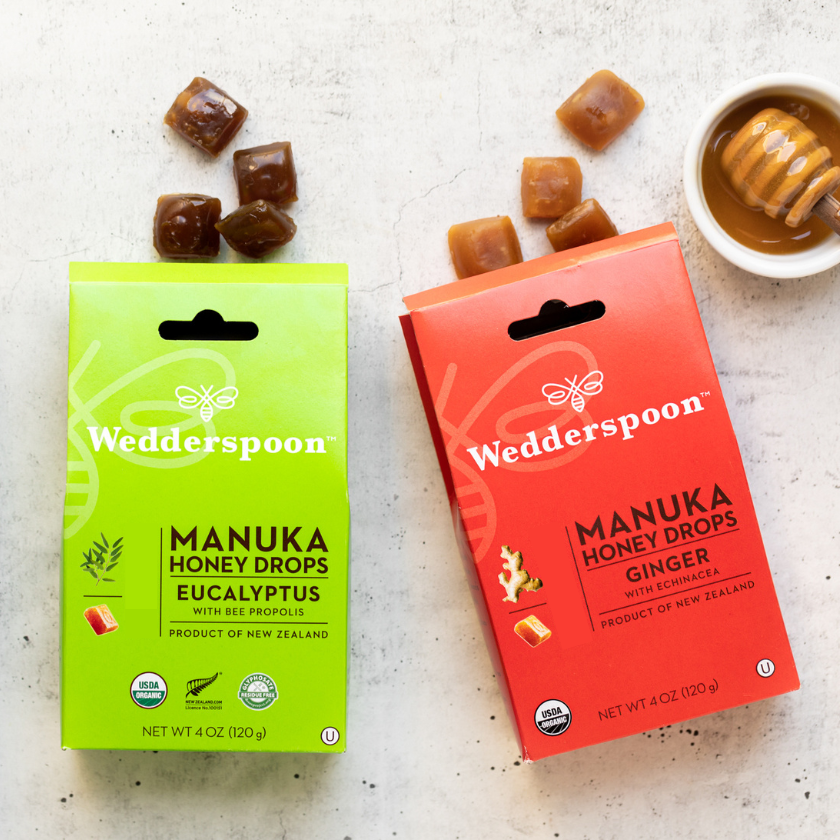 Two boxes of Wedderspoon Manuka Honey Drops on a light surface with honey and a dipper.