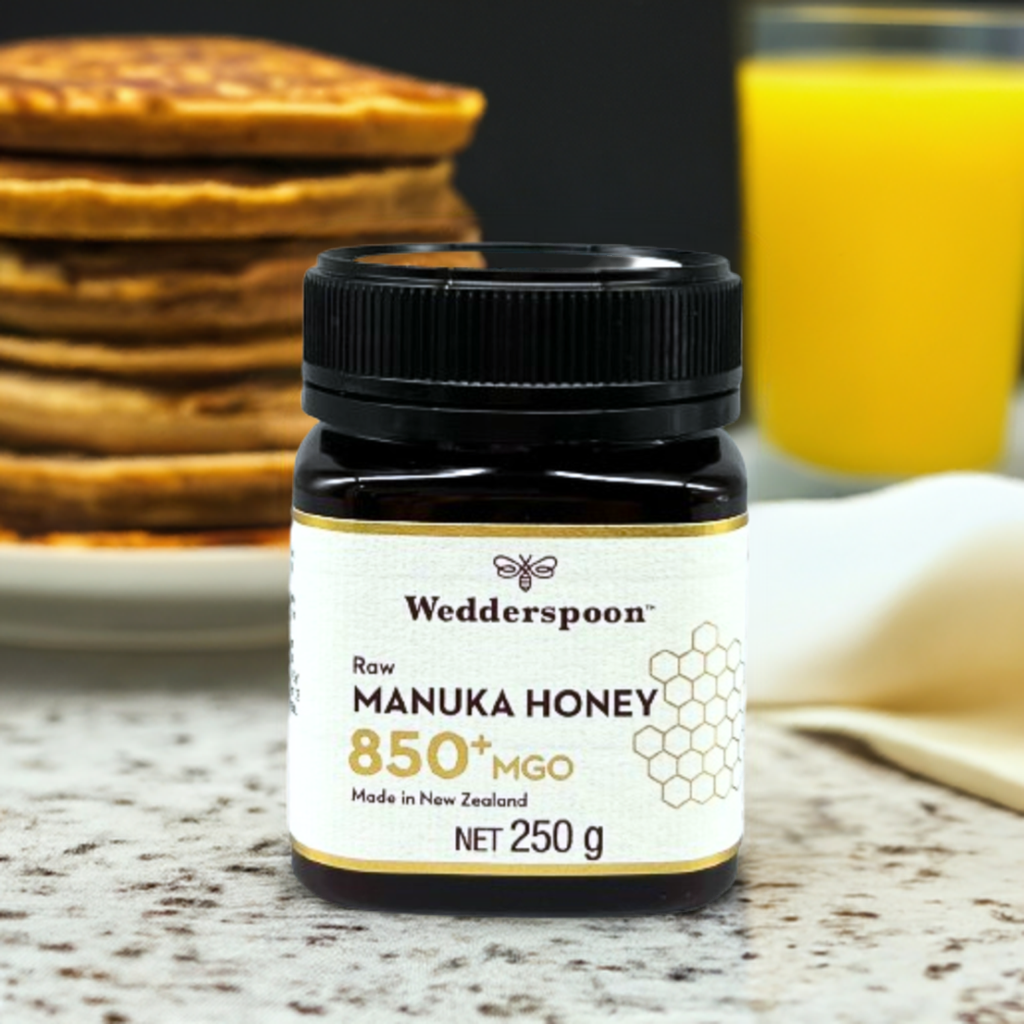 Jar of Wedderspoon raw manuka honey 850 mgo 250g on kitchen table with pancakes and orange juice