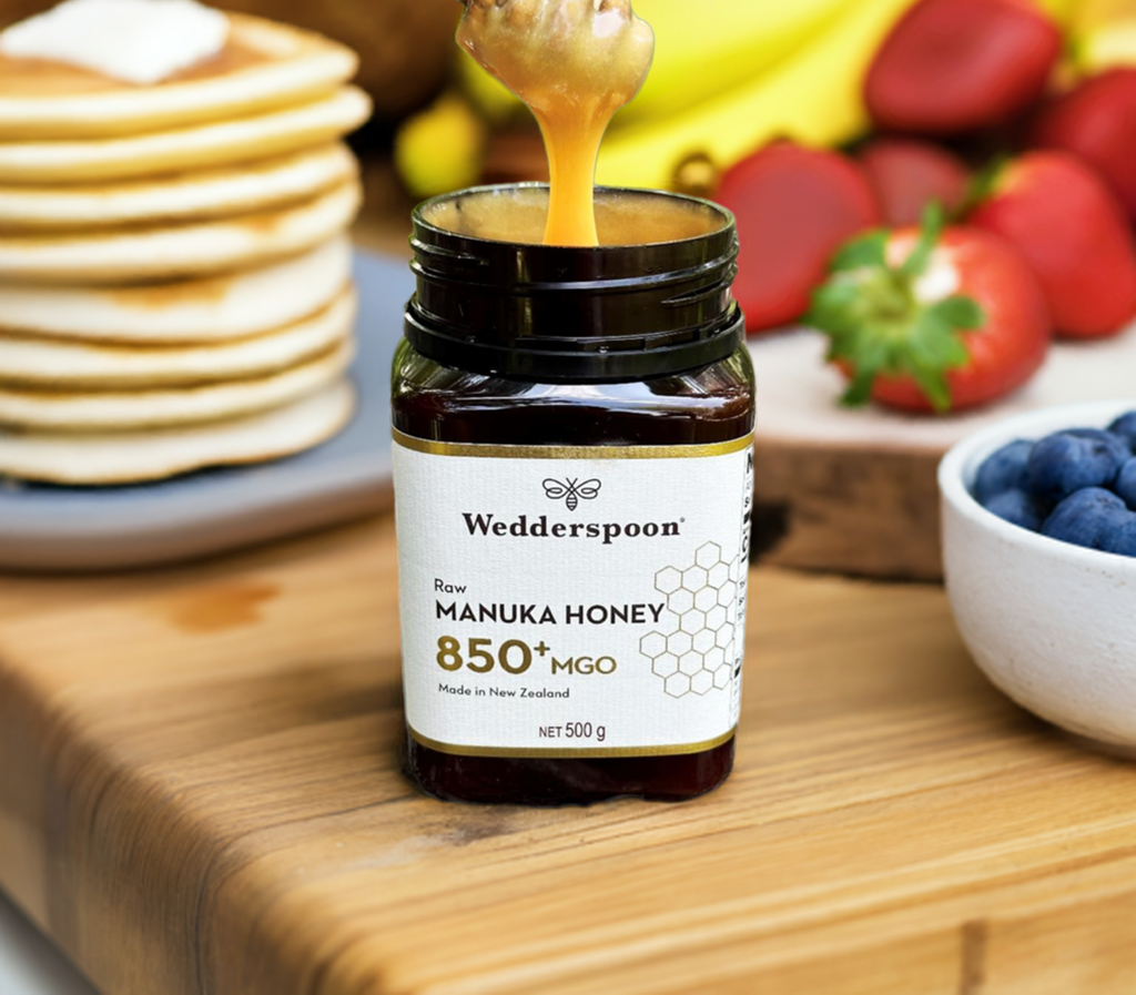 Wedderspoon raw manuka honey 850 mgo jar with honey dripping on wooden table next to pancakes and fresh fruit