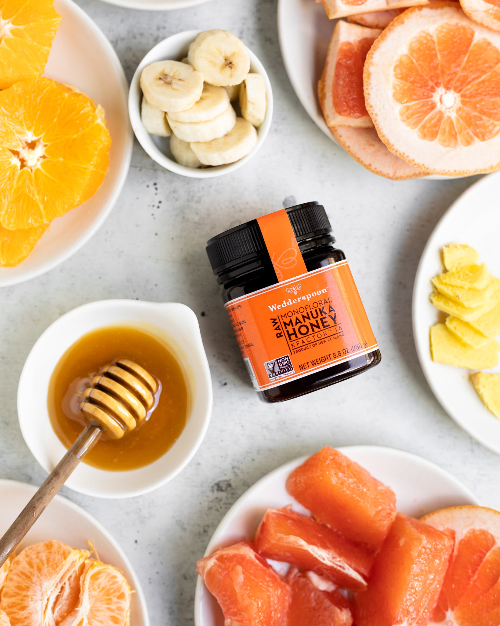 Wedderspoon raw Manuka honey KFactor 16+ jar on table with honey dipper and sliced citrus and bananas