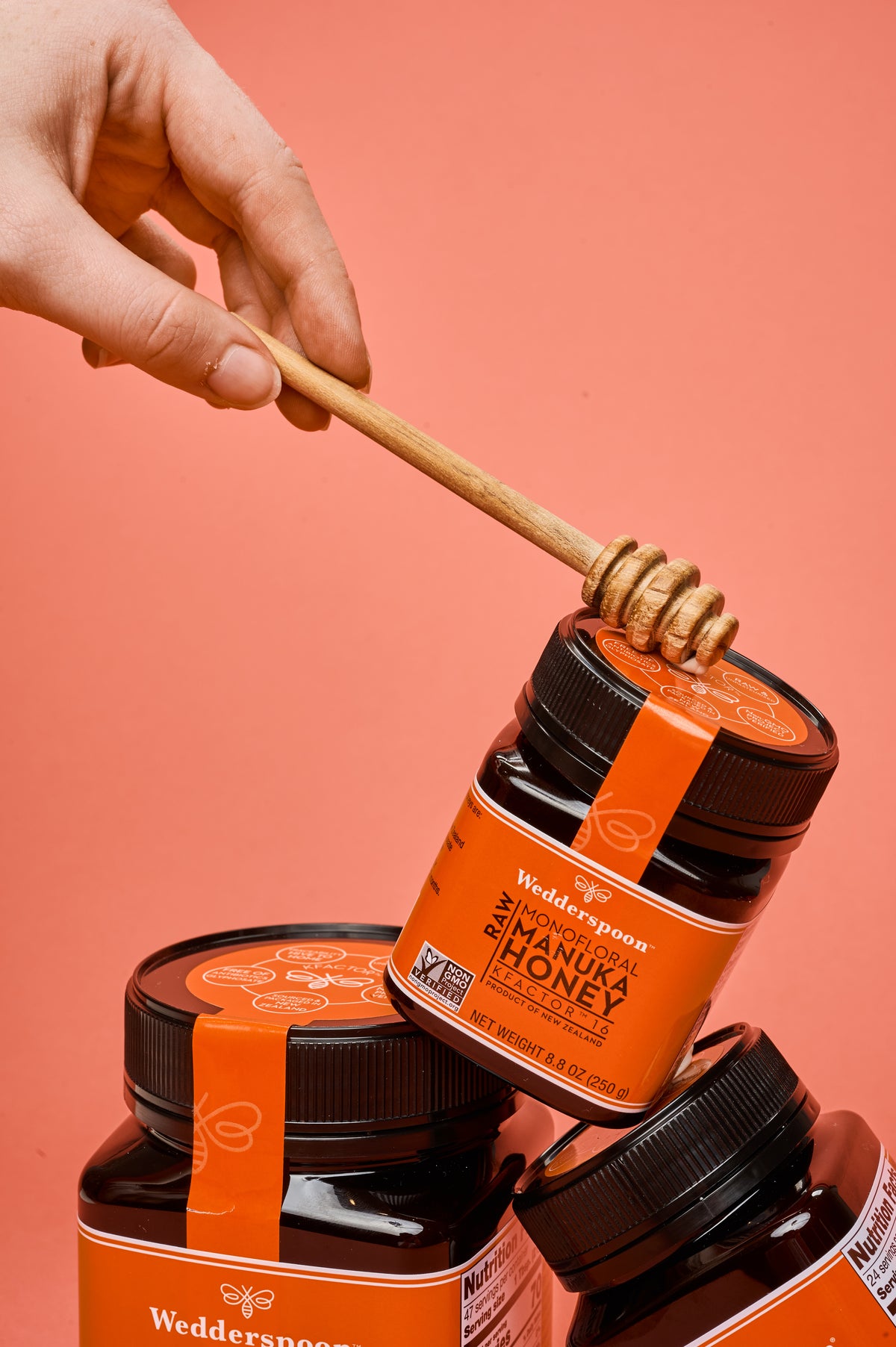 Hand with honey dipper over stacked Wedderspoon jars of Wedderspoon raw Manuka honey KFactor 16+ with orange labels