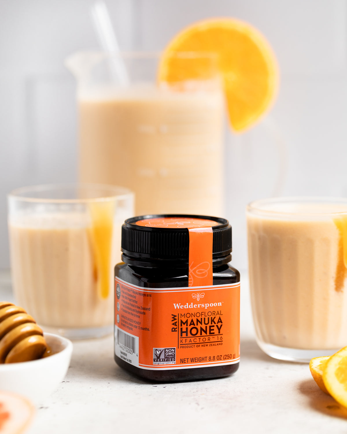 Jar of Wedderspoon raw Manuka honey KFactor 16+ with orange smoothies and honey dipper