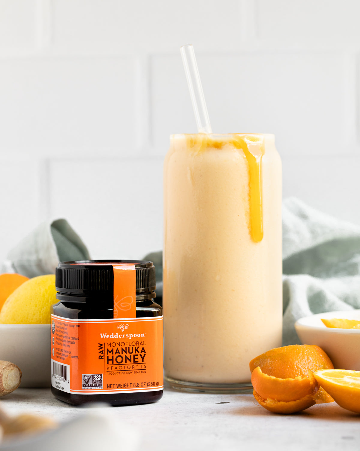 Wedderspoon raw Manuka honey KFactor 16+ jar beside citrus smoothie with honey drizzle